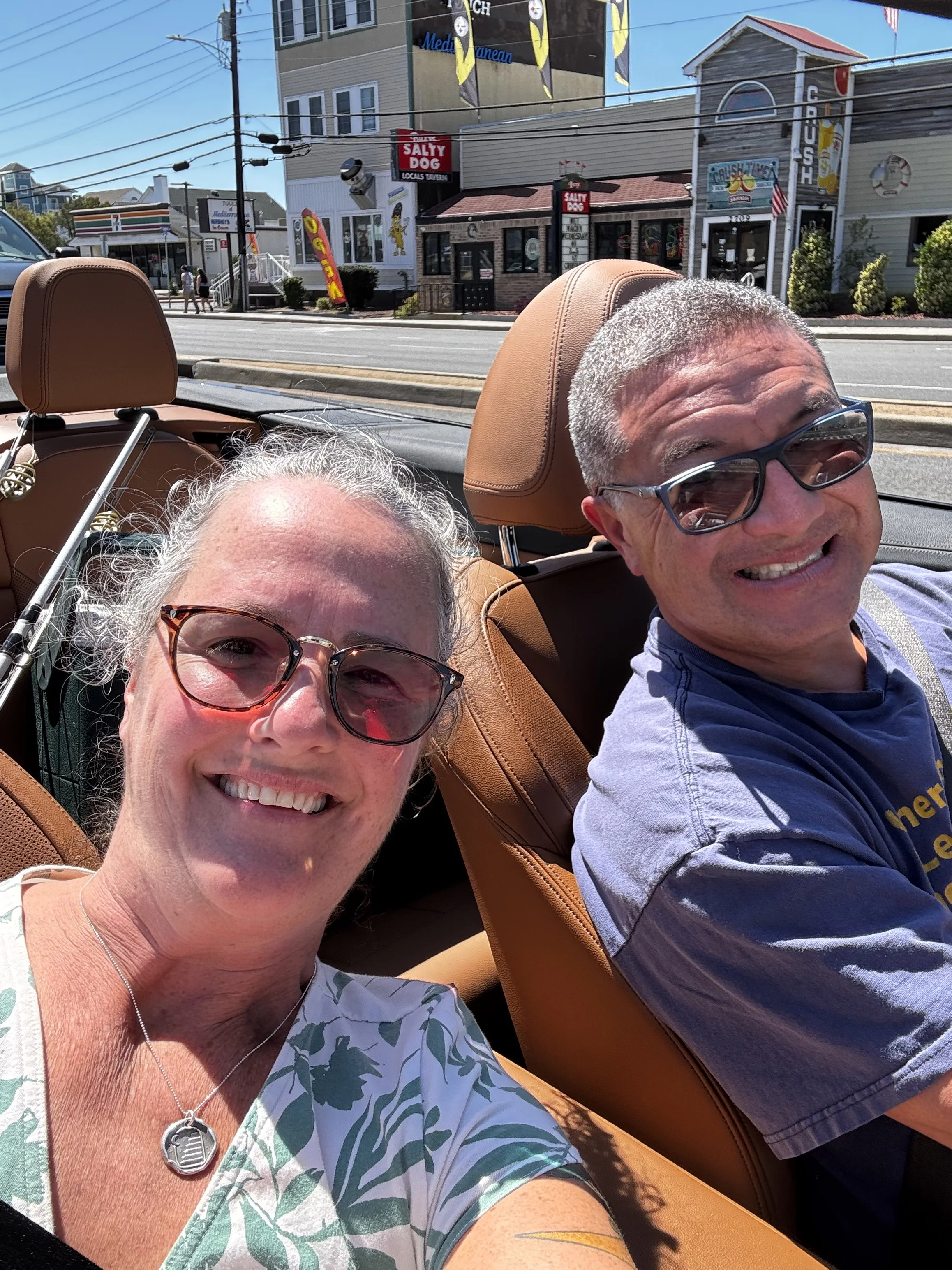 Two smiling people taking a selfie in a convertible car with a city street background, including buildings and signage for businesses like Salty Dog and a 7-Eleven.