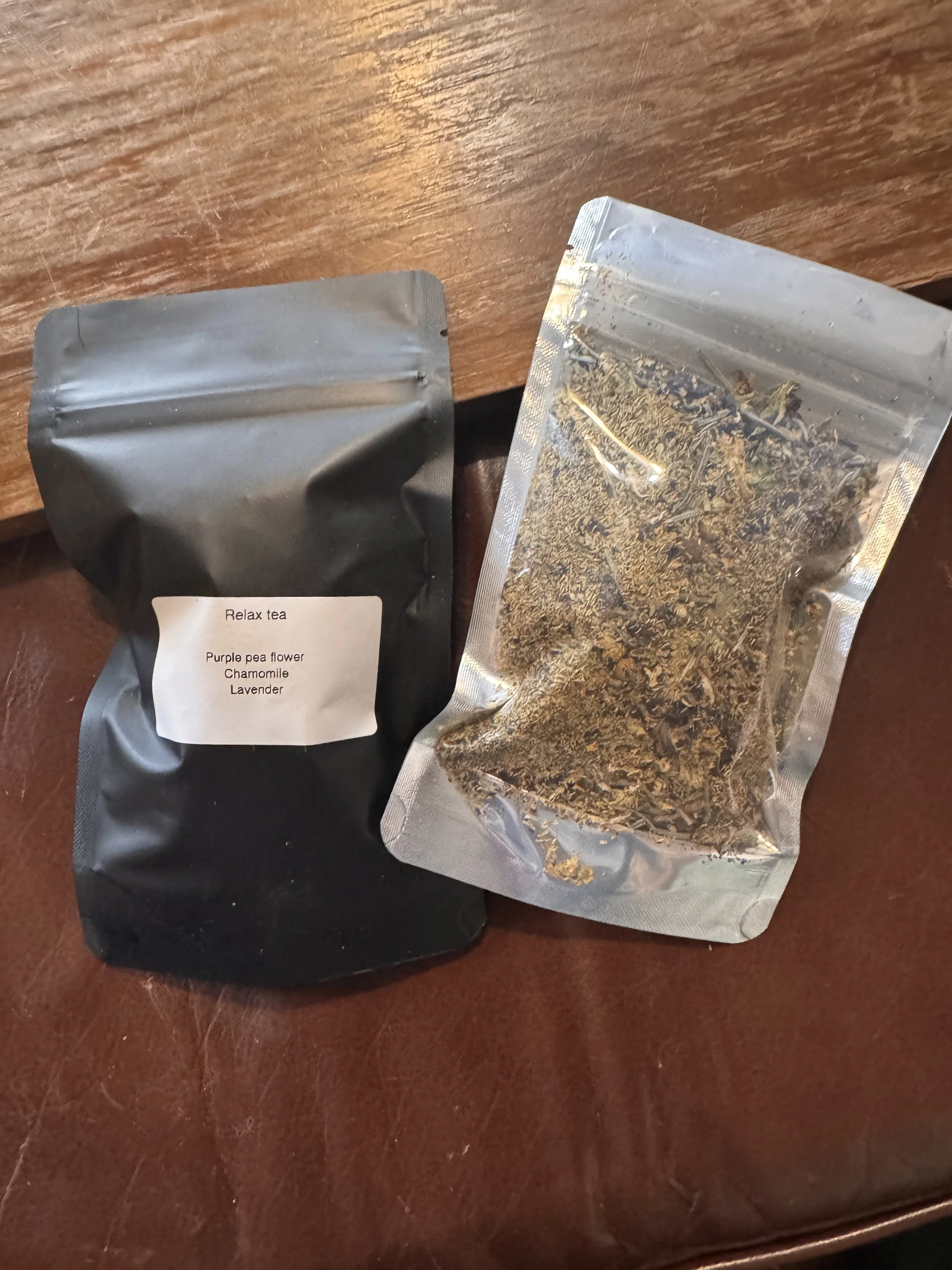 Two packages of tea on a wooden surface, one labeled "Relax tea" with ingredients including purple pea flower, chamomile, and lavender, and the other containing loose dried herbs.