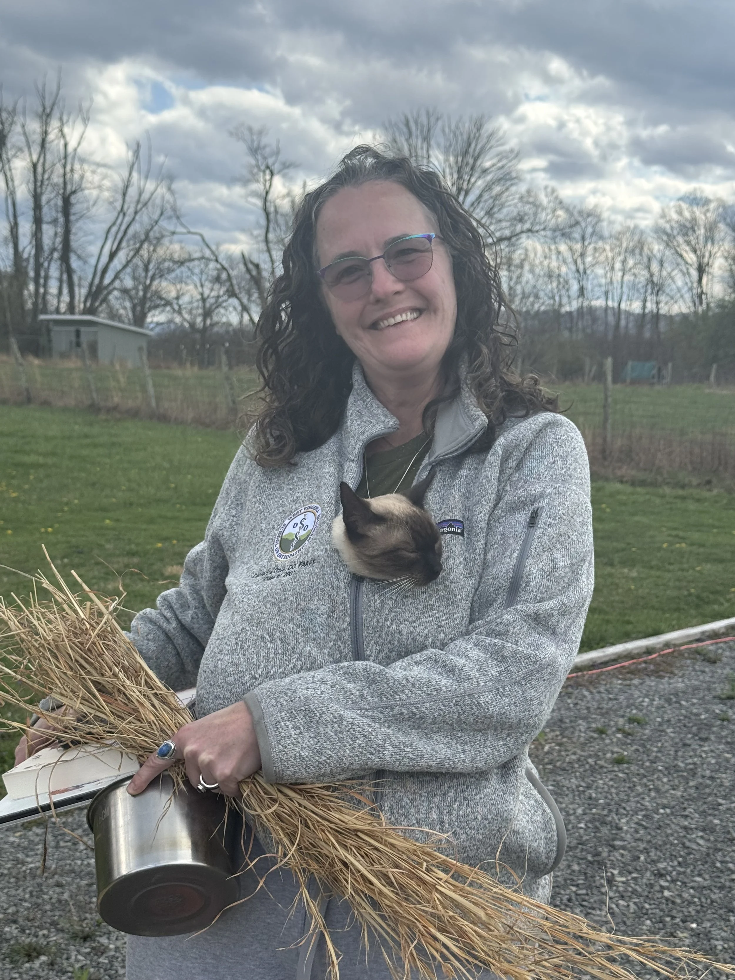A woman holding a bundle of dried grass with a siamese cat resting in her jacket. She is outdoors with trees and a cloudy sky in the background.
