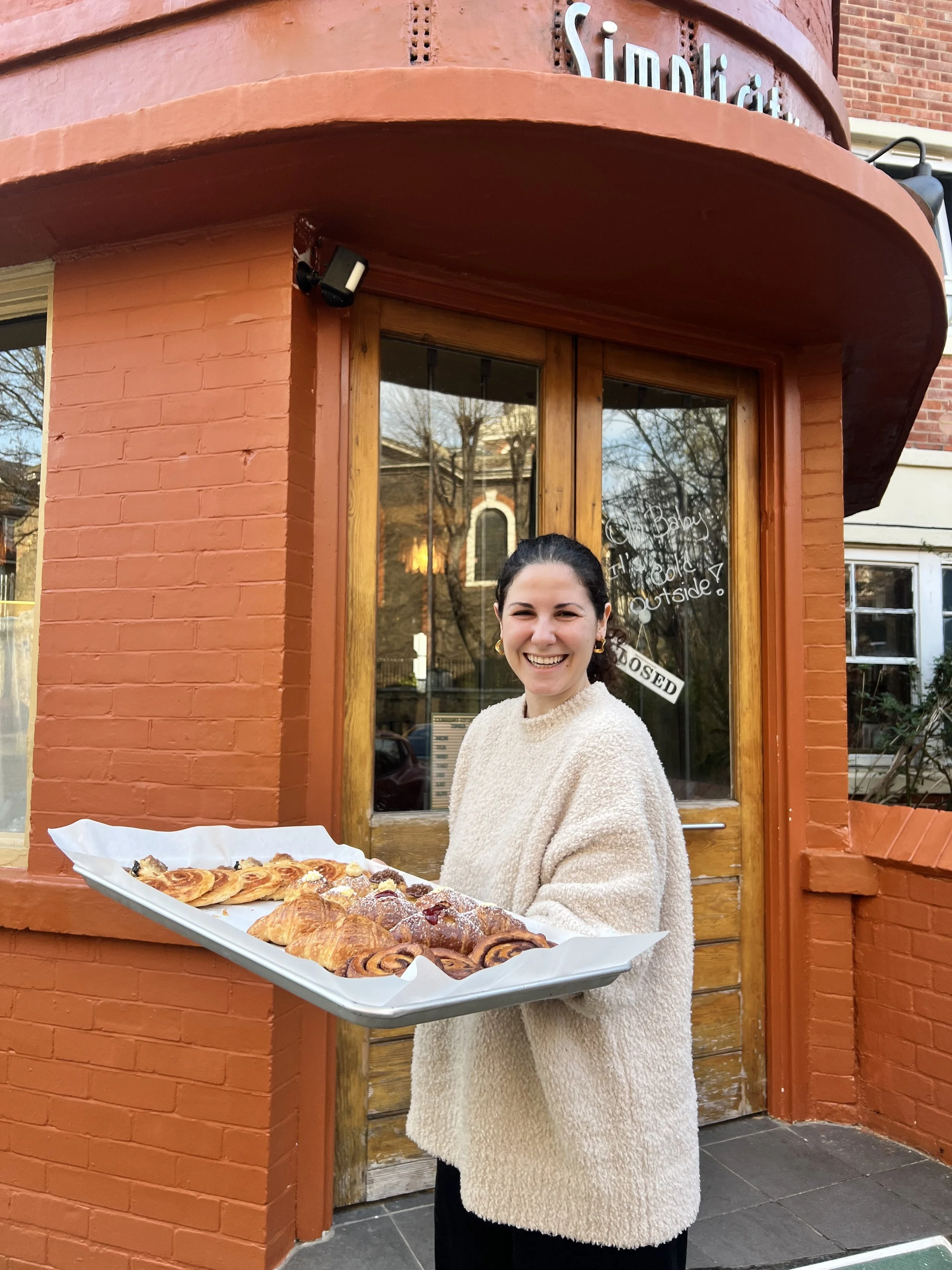 A woman smiling and holding a large tray of assorted baked goods outside a café with red brick walls and a window.