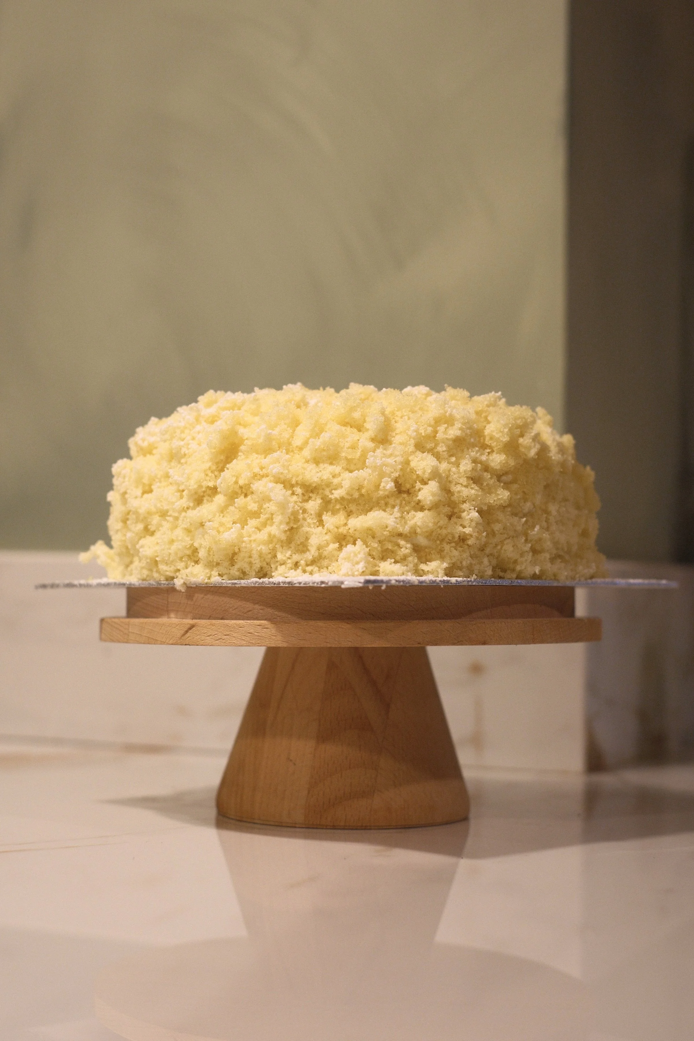 A round, yellow cake with crumbly texture on a wooden cake stand.