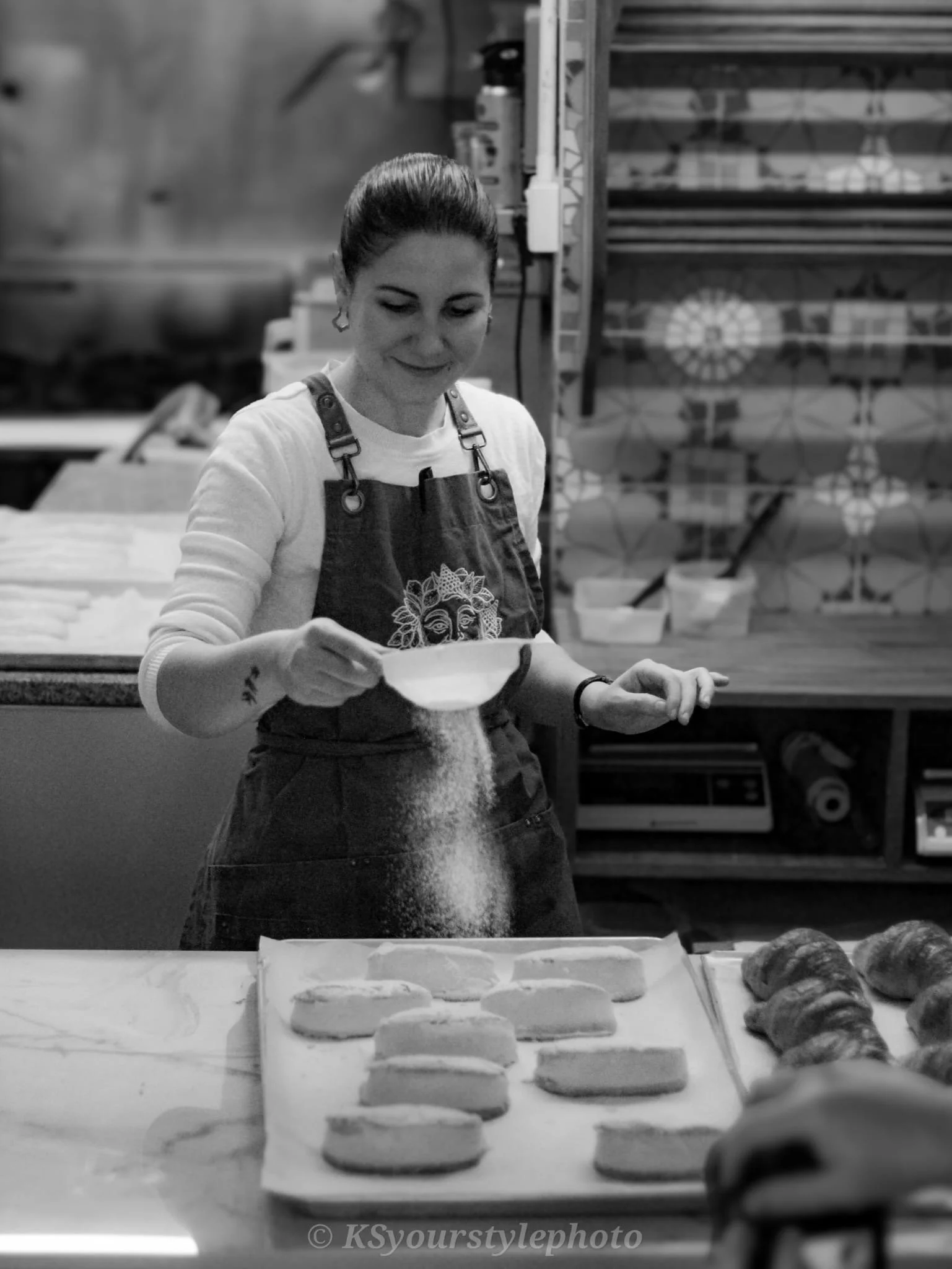 A woman wearing an apron sprinkles flour onto a countertop while baking bread or pastries, with rows of dough in front of her and baking ingredients on shelves in the background.
