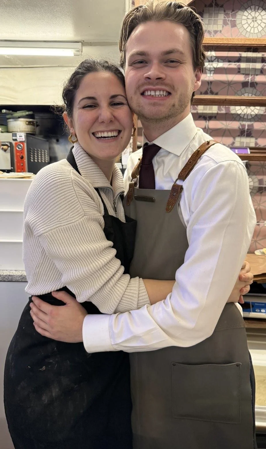 A woman and man hug and smile in a kitchen or cafe setting, both wearing aprons.