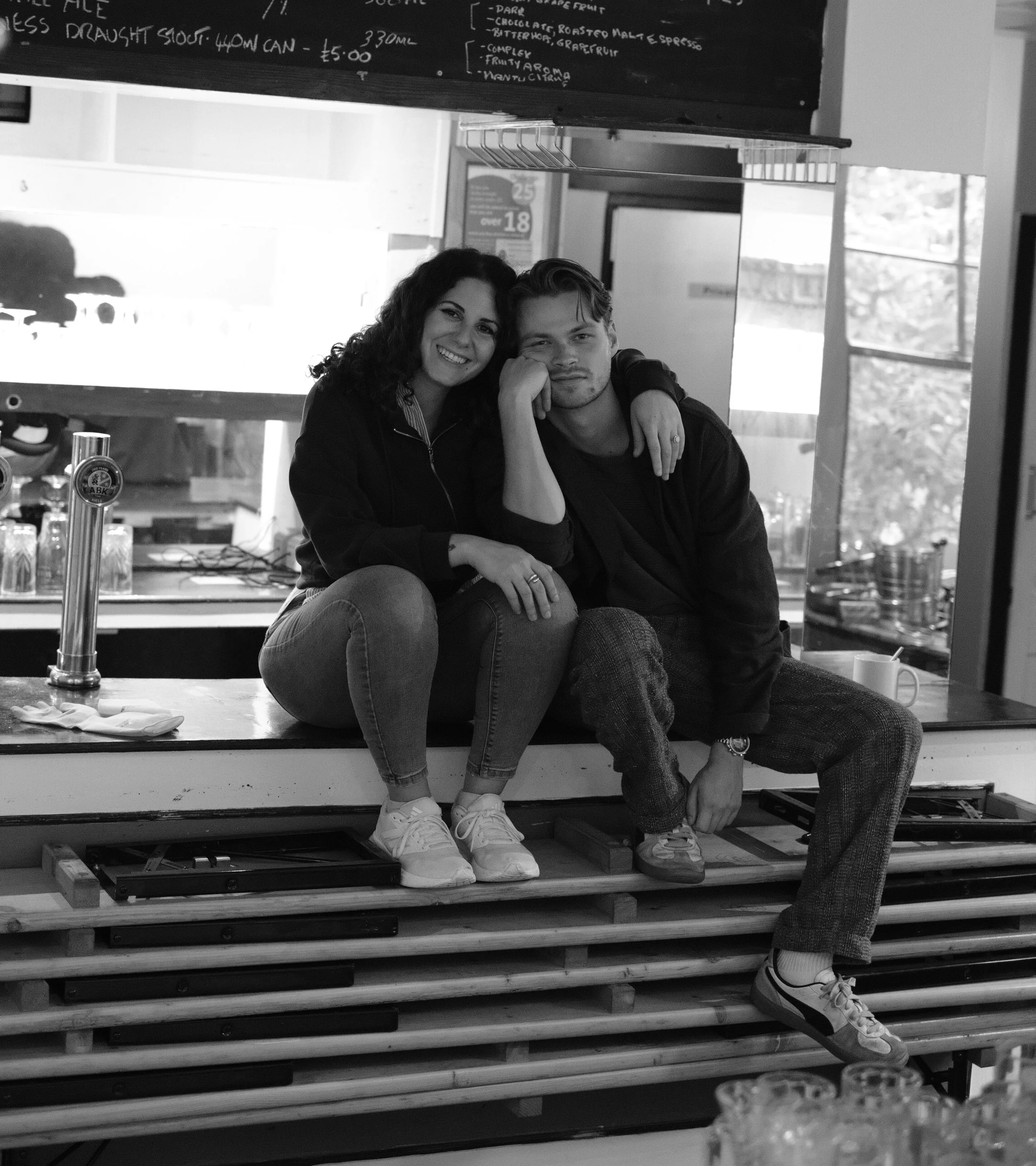 Black and white photo of a smiling woman and a man sitting on a platform, their arms around each other's shoulders. The woman has curly hair and is wearing a dark jacket and sneakers; the man has wavy hair and is wearing a dark jacket and patterned pants. Behind them, a window lets in natural light and a chalkboard menu is visible at the top of the image.