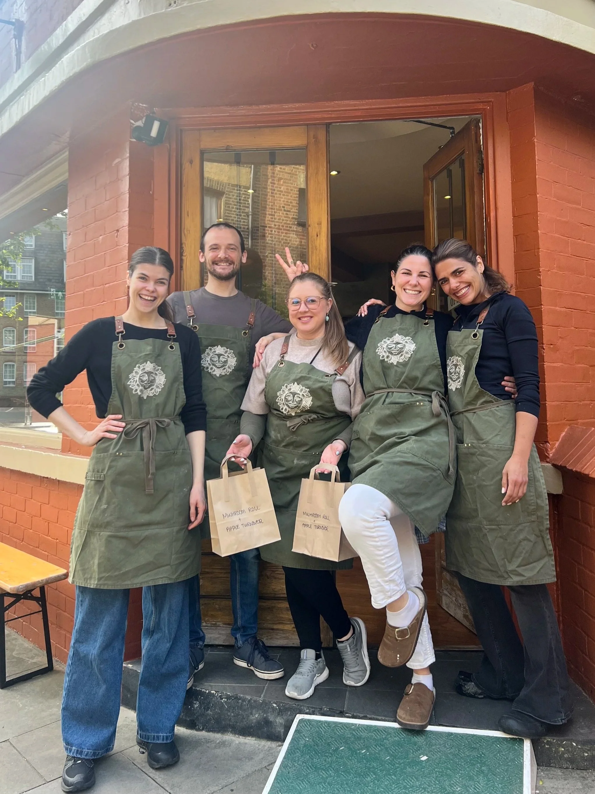 Five people standing outside a restaurant or café, smiling and posing for the photo. They are wearing matching green aprons with white embroidered designs. Two of them are holding brown paper bags labeled with menu items, including mushroom roll and apple turnover. The group appears to be staff or volunteers.
