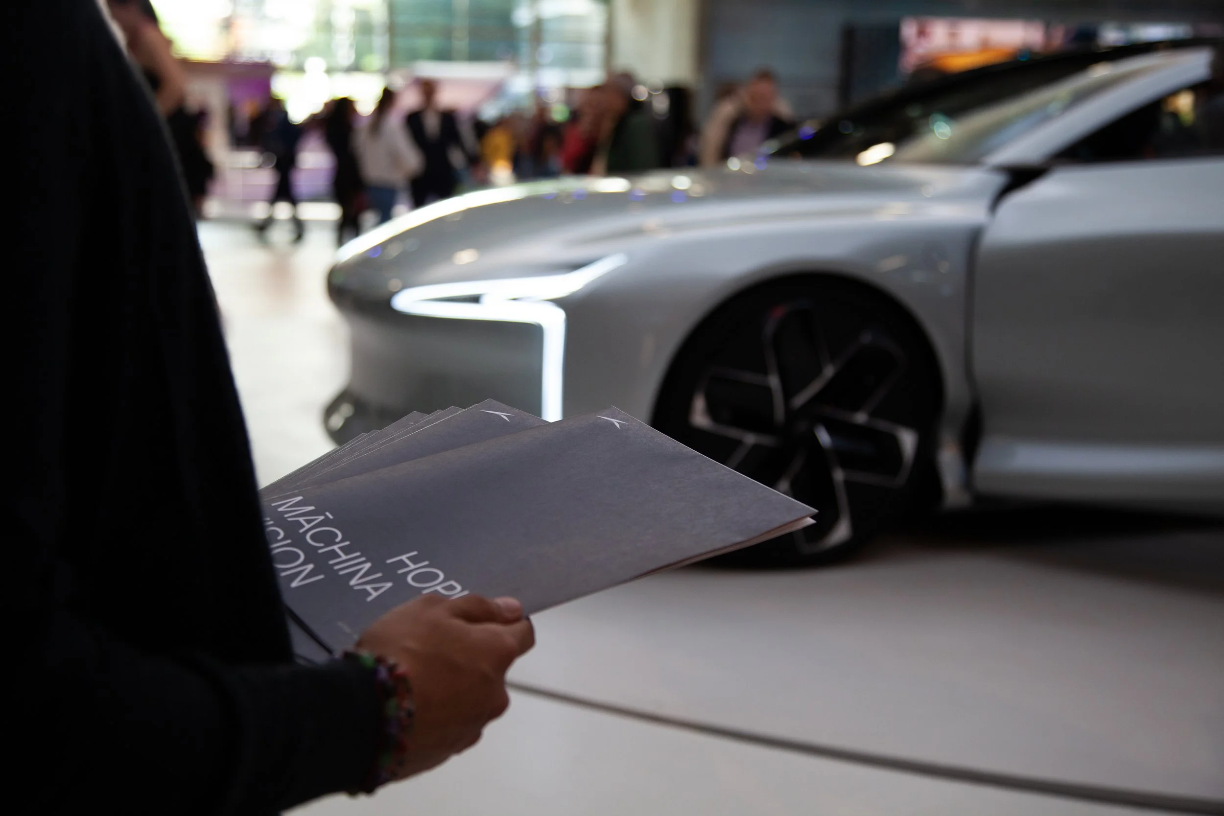 Une personne tenant des brochures ou catalogues devant une voiture de luxe dans un salon automobile, avec plusieurs personnes en arrière-plan.