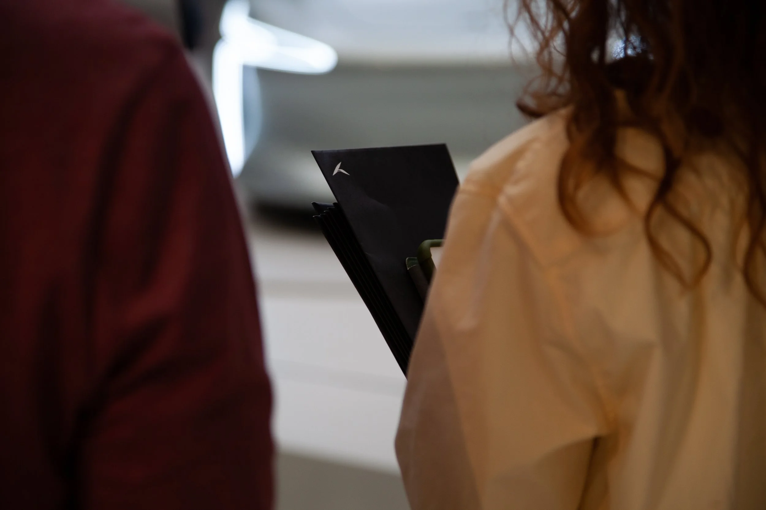 Personnes debout près de voitures en arrière-plan, l'une d'elles tenant un bouquet de documents ou de brochures.