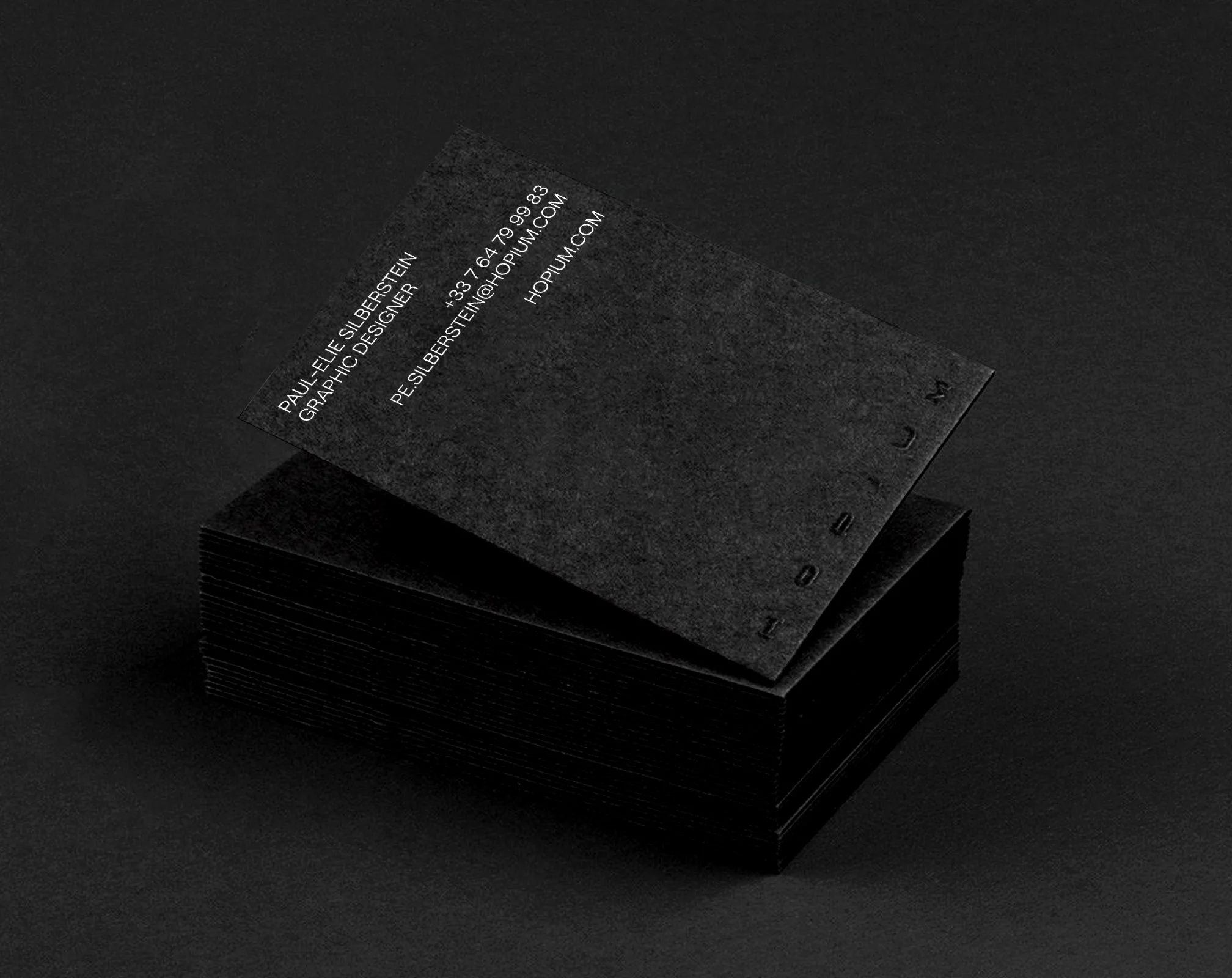 Piles de cartes de visite noires avec texte blanc, dont une carte visible montrant les coordonnées et le nom d'un graphiste.