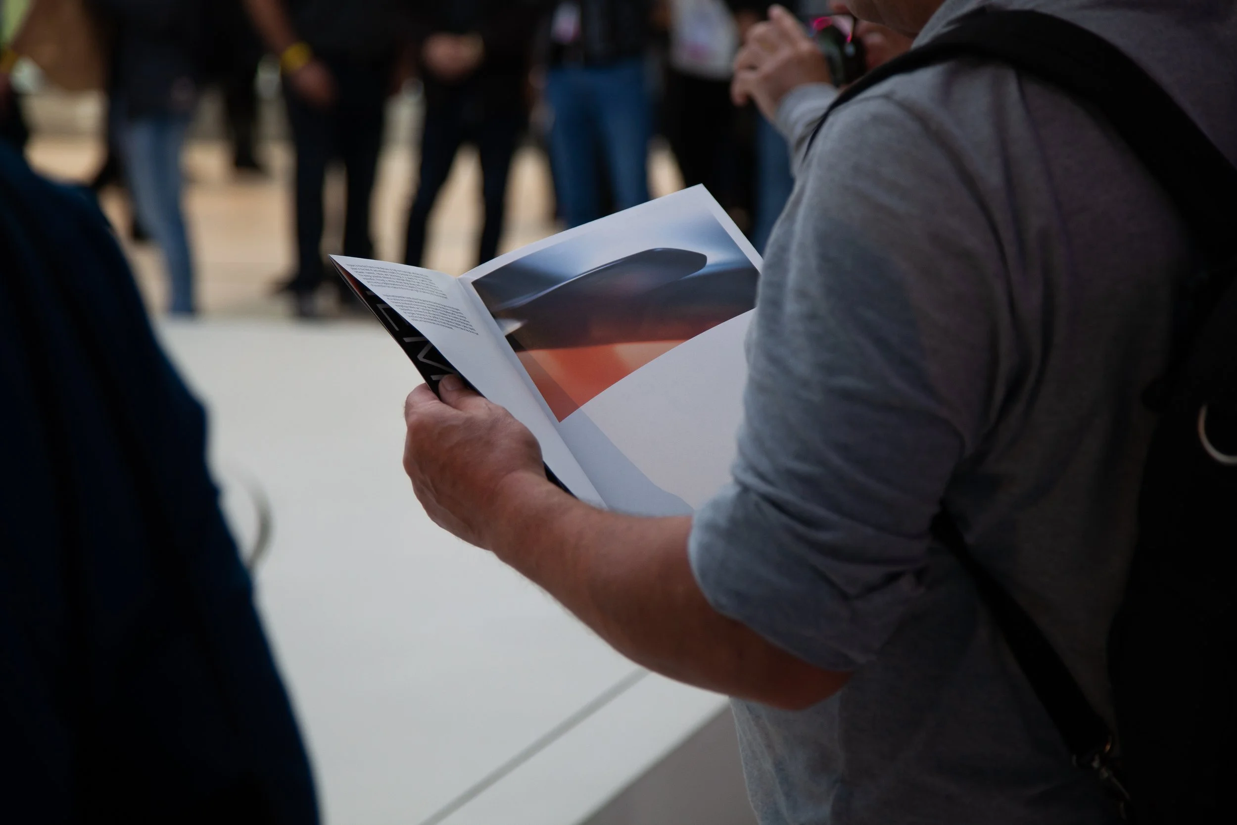 Personne tenant un catalogue d'exposition dans une galerie d'art, avec un groupe de personnes en arrière-plan.