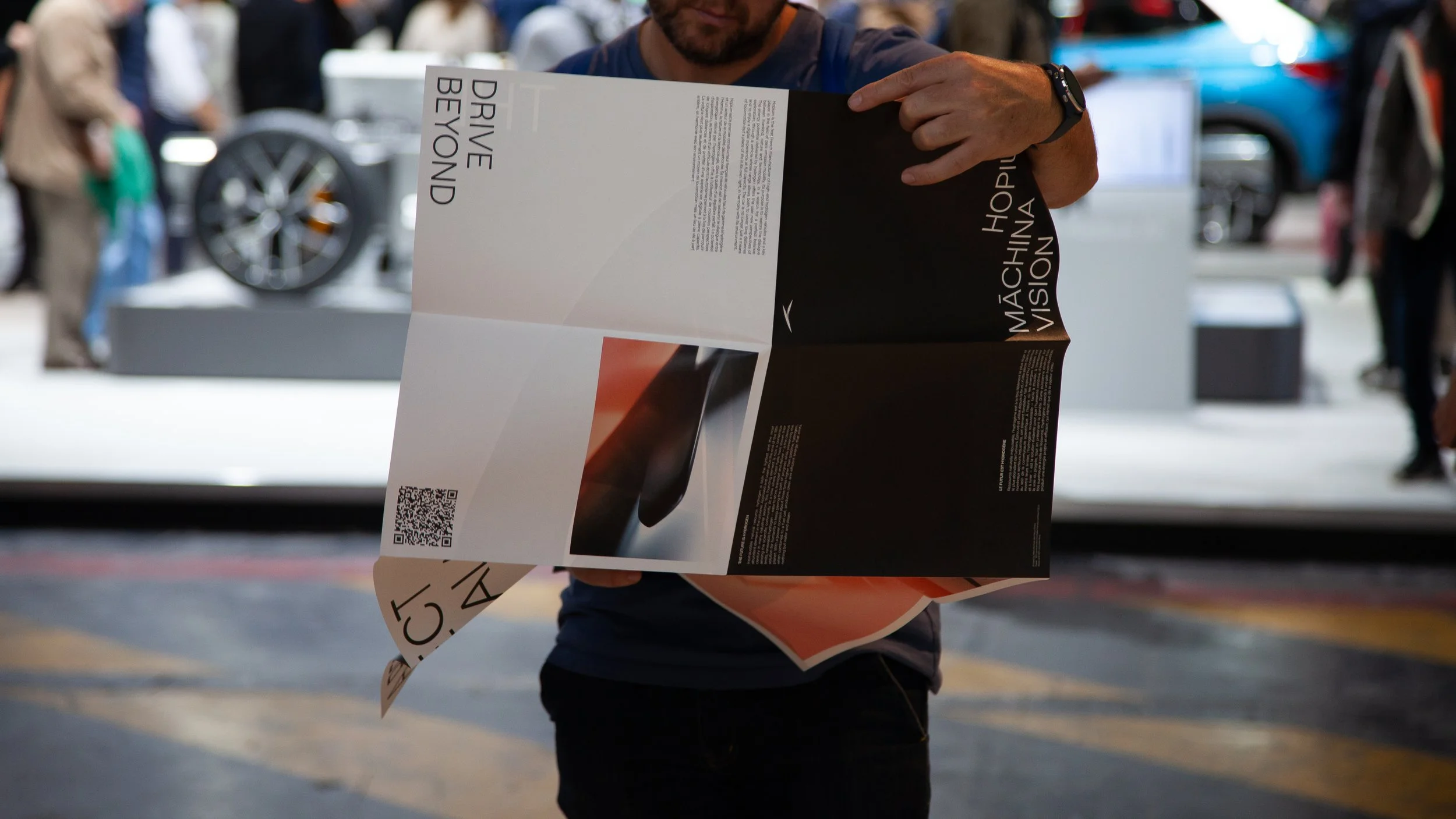 Une personne tenant un dépliant présentant une tablette ou un appareil électronique avec du texte en anglais et un code QR, dans un environnement d'exposition ou de foire.