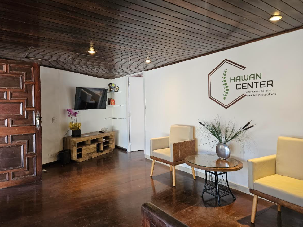 Interior of a wellness center reception area with two cream-colored chairs, a small round wooden table with a vase and plant, a wall-mounted TV, shelves with decorative items, and a logo on the wall reading "Hawan Center," featuring a DNA strand and hexagon outline.