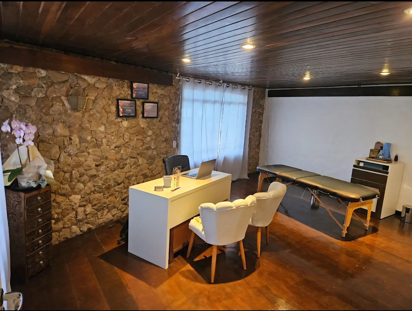 Interior of a room with a stone accent wall, a white desk with a laptop, two white chairs, a massage table, and a small white cabinet. The room has dim lighting and a window with white curtains.
