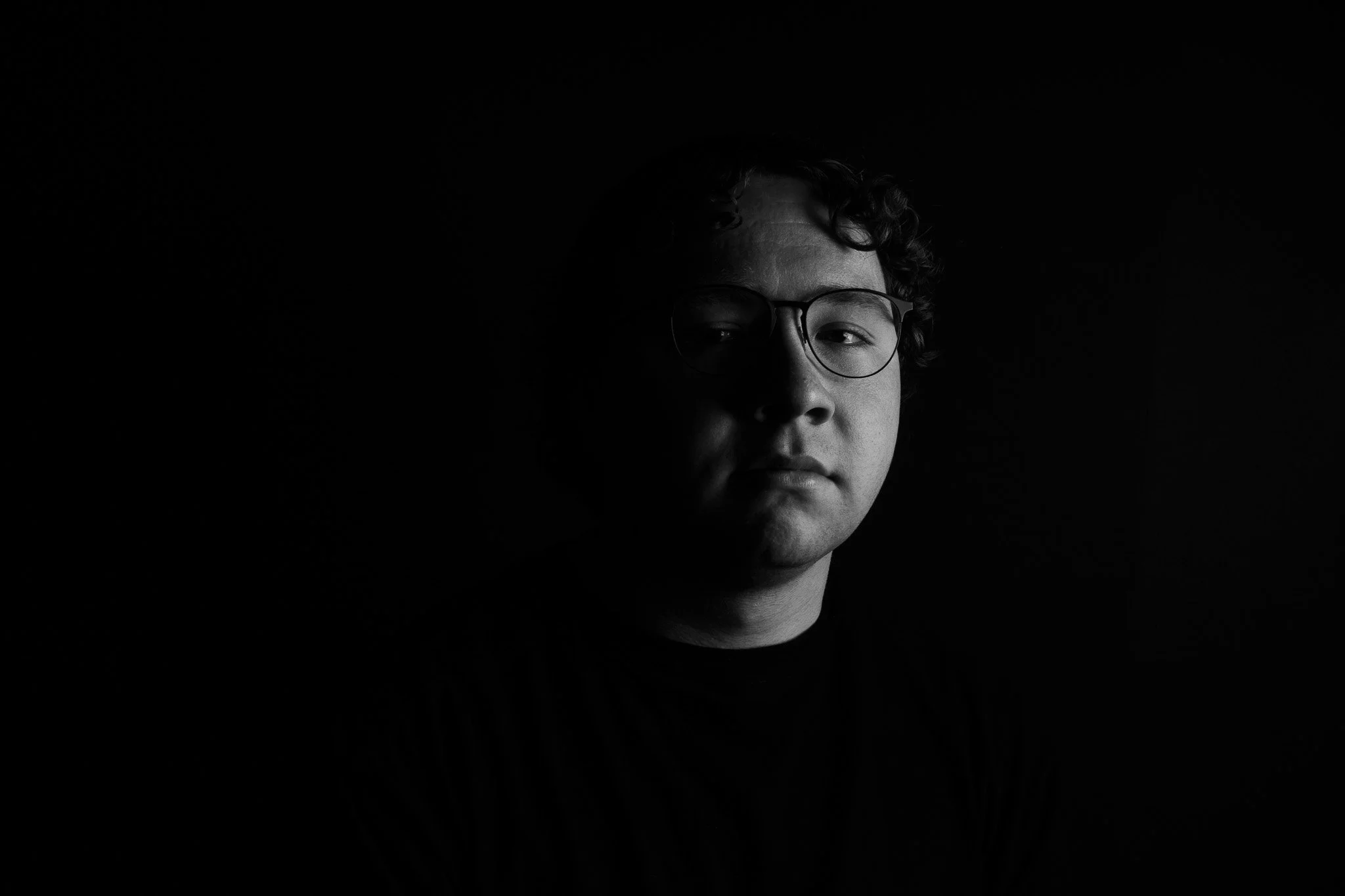 Black and white photo of a young person with glasses, partly obscured by darkness.