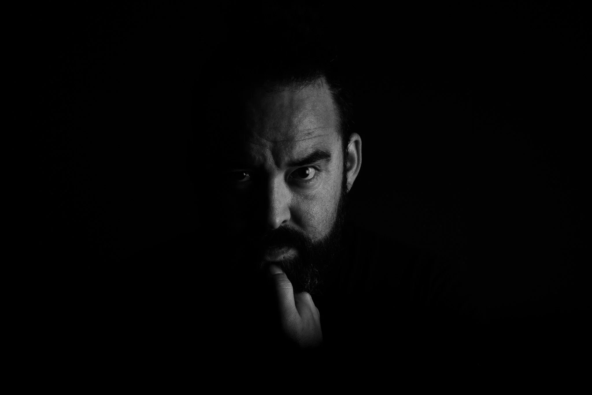 Black and white photo of a man with dark hair and beard, partially visible, making a gesture with his finger near his lips in a contemplative pose.