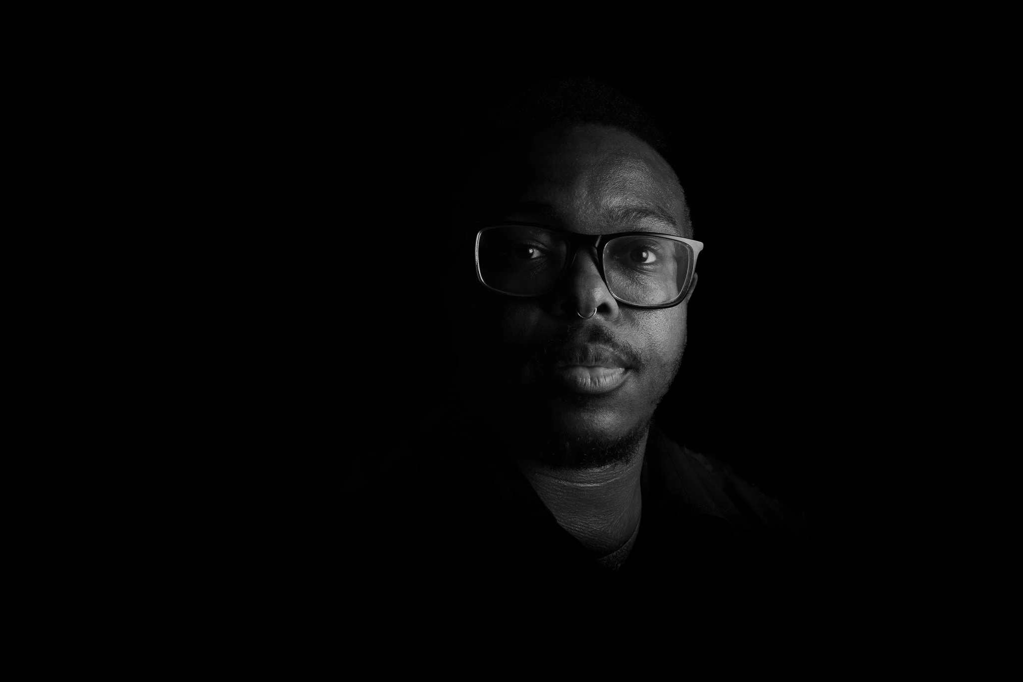 Black and white portrait of a man wearing glasses with a serious expression, dark background, lighting focused on his face.
