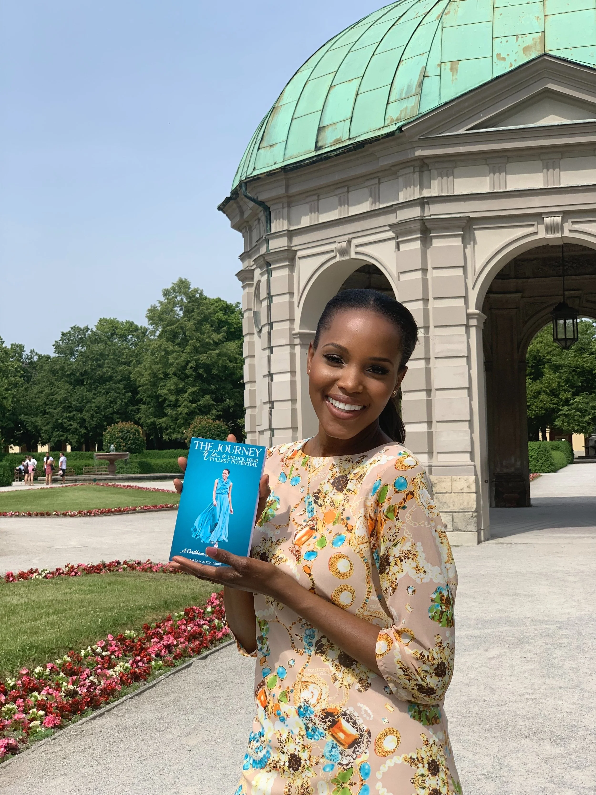 A smiling woman holding a brochure with a blue dress on the cover outdoors near a historic building with a green dome and arches, with a garden and trees in the background.