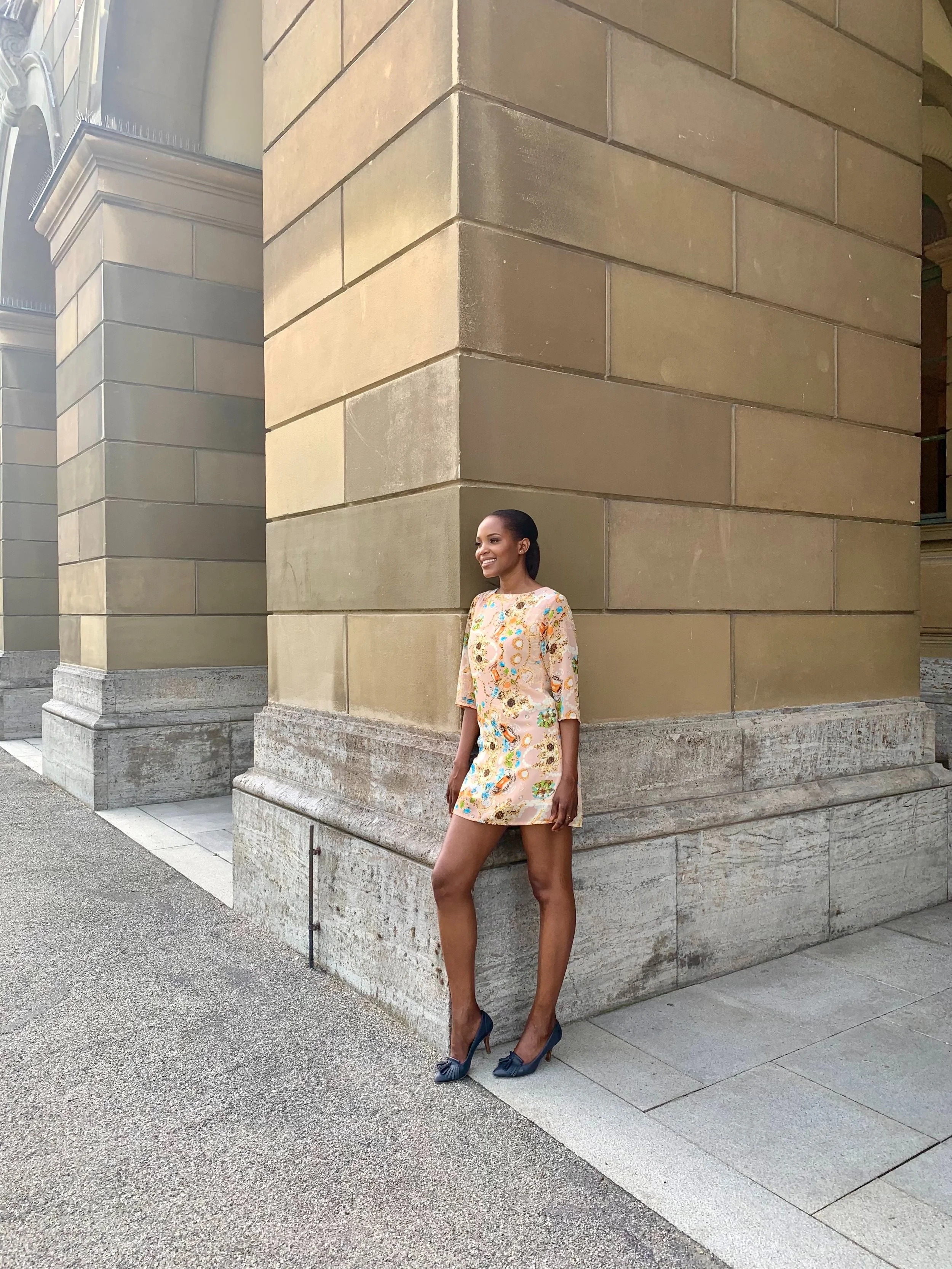A woman in a light-colored floral dress and high heels standing against a large stone building exterior.