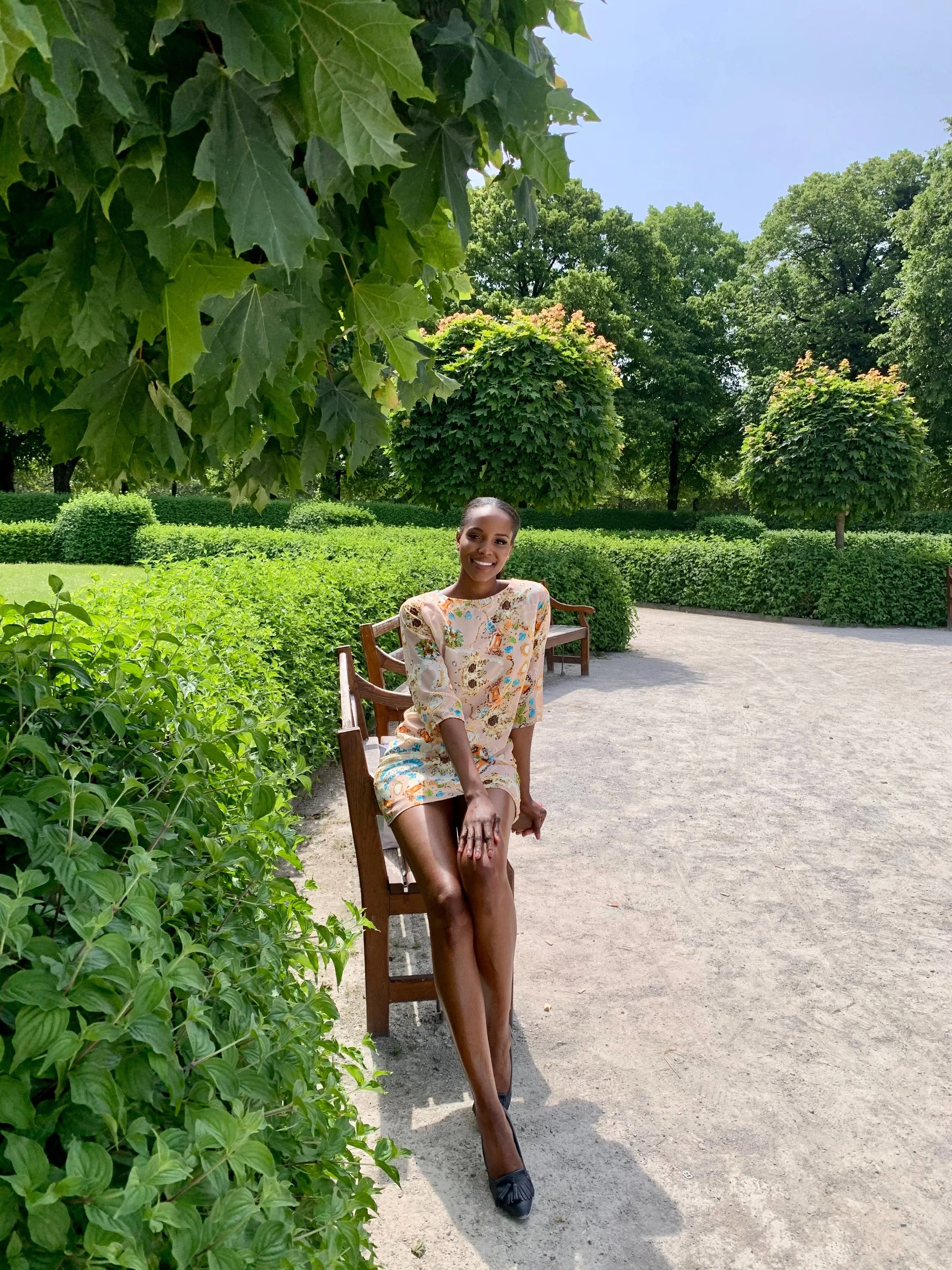 A woman sitting on a wooden bench in a lush green park, smiling. She is wearing a light-colored, floral dress and black shoes. There are trees and bushes in the background under a blue sky.