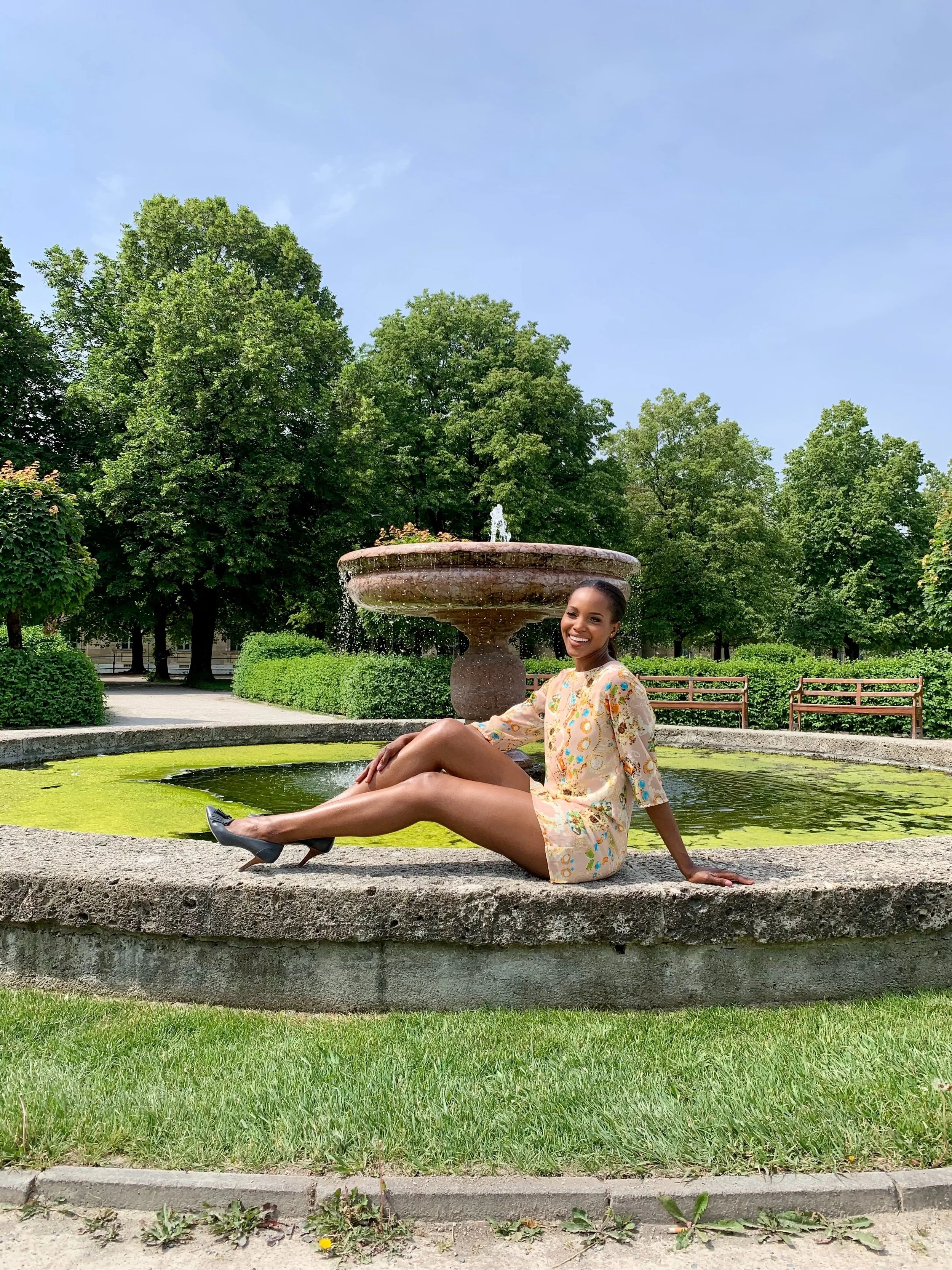 Woman sitting on the edge of a fountain in a park, smiling, wearing a patterned dress and high heels, with green trees and blue sky in the background.