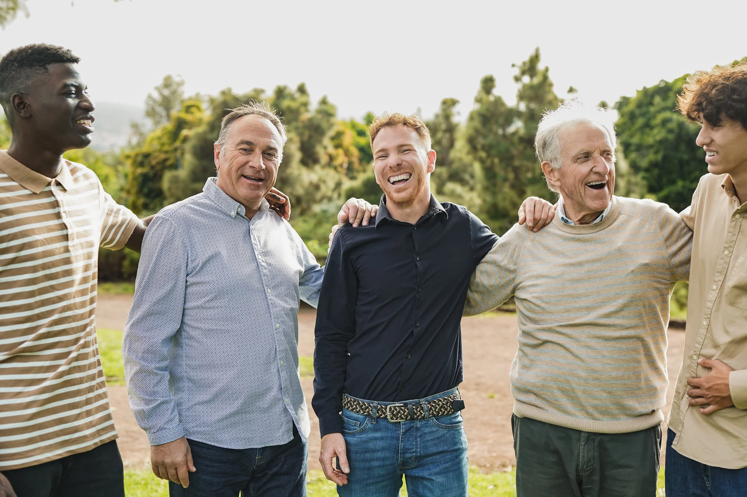 Group of six diverse men standing outdoors with arms around each other, smiling, in a park with trees in the background.