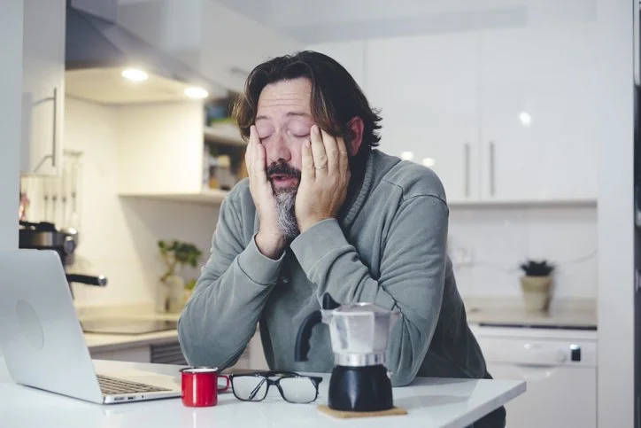 A man with dark hair and a beard sitting at a kitchen counter, looking frustrated or exhausted with his hands on his face. There are a laptop, a mug, and a pair of glasses in front of him.