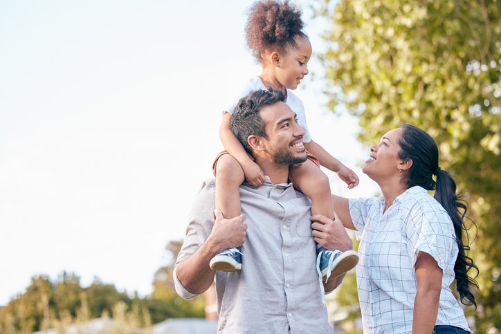A happy family of three outdoors; a man carries a young girl on his shoulders, and a woman looks at them smiling.