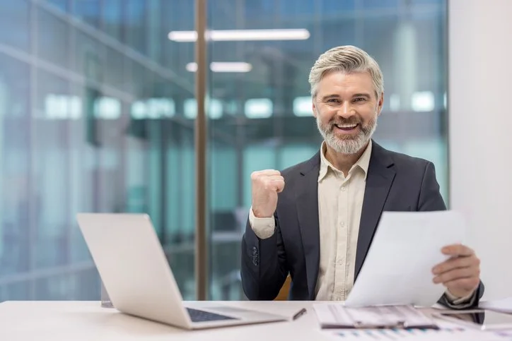 An older man in a business suit with gray hair and a beard, smiling and celebrating with a clenched fist, sitting at a desk with a laptop and papers in a modern office with glass walls.