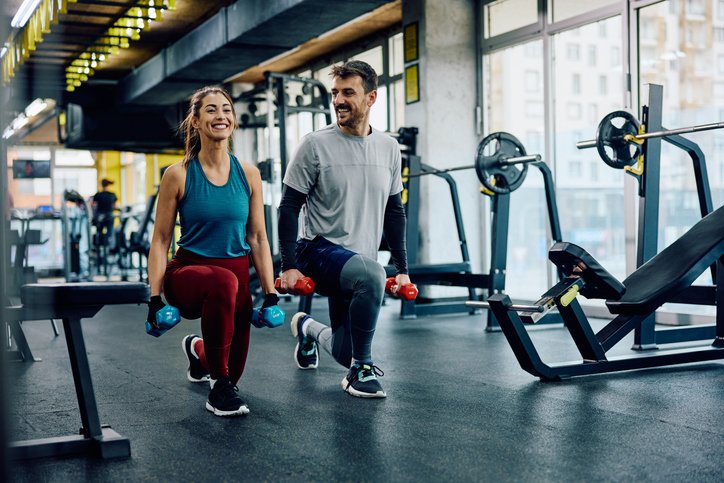 A man and woman at the gym doing lunges with dumbbells, smiling at each other, with fitness equipment and large windows in the background.