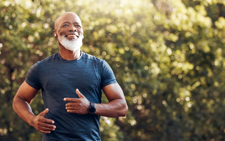 An older man with a beard and a bald head, wearing a blue athletic shirt and a black watch, smiling and jogging outdoors in a park with trees and sunlight in the background.