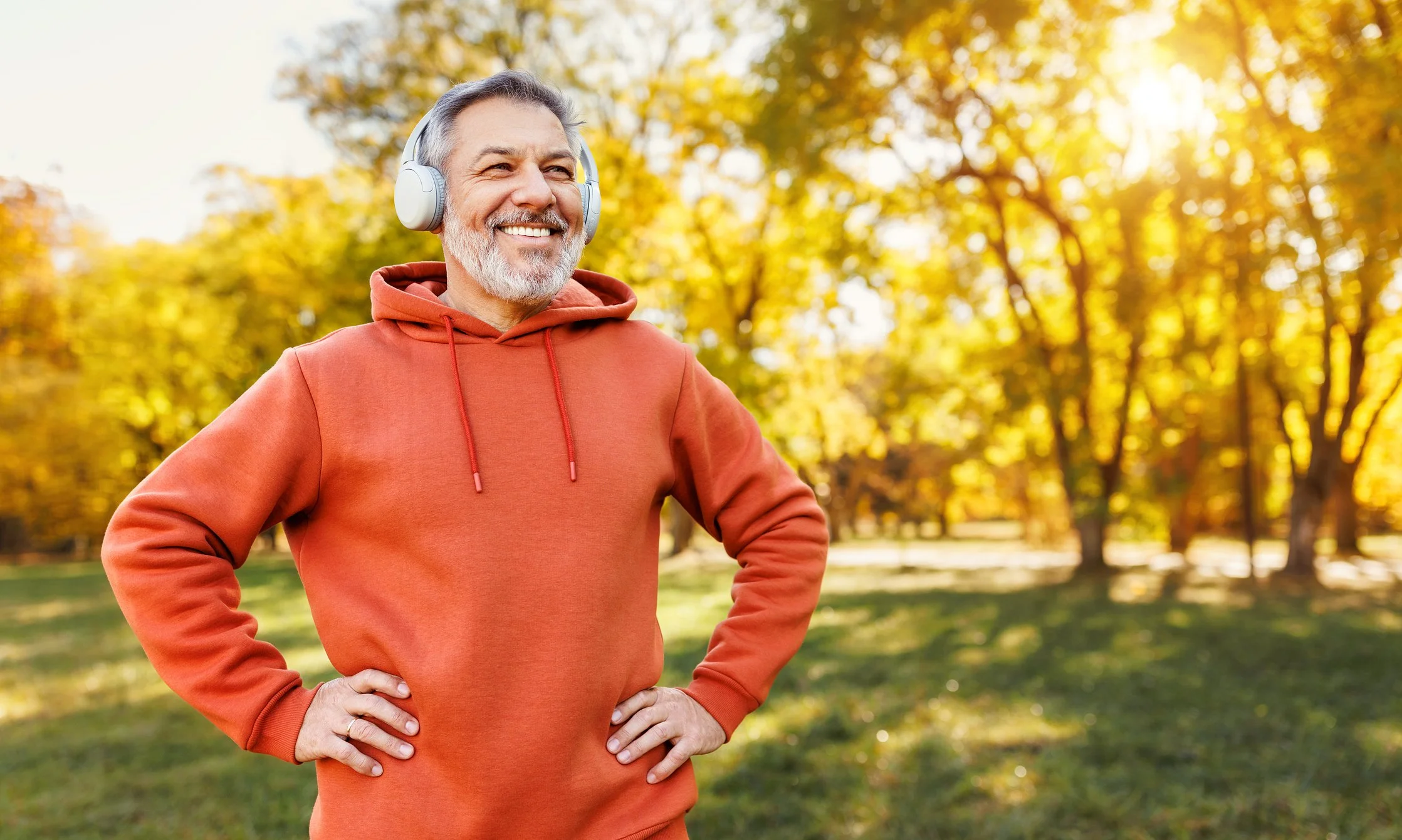 An older man smiling outdoors in a park during autumn, wearing a rust-colored hoodie and headphones.