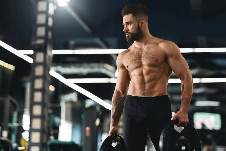 A muscular man with a beard and short dark hair lifting weights in a gym.