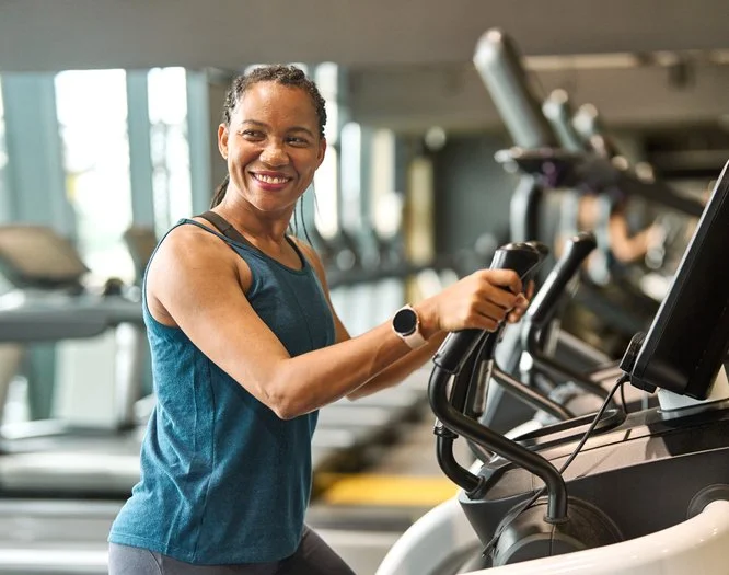 A woman smiling and working out on an elliptical machine in a gym.