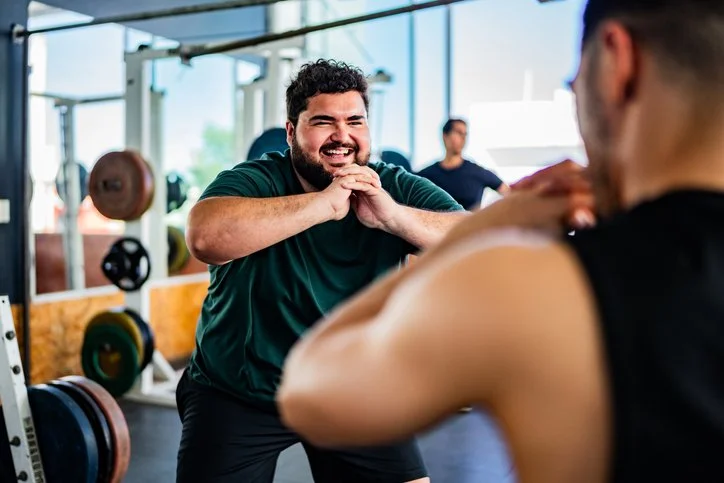 Two men in a gym, one smiling with hands clasped in front of him, the other with back facing the camera, working out.