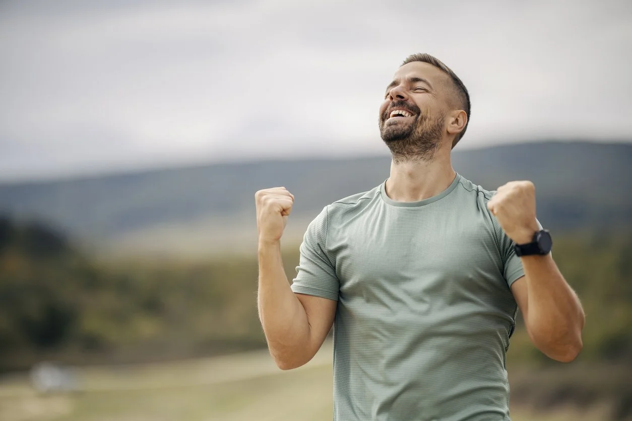 A man celebrating outdoors with clenched fists, smiling, wearing a gray athletic shirt and smartwatch, with a blurred natural landscape in the background.