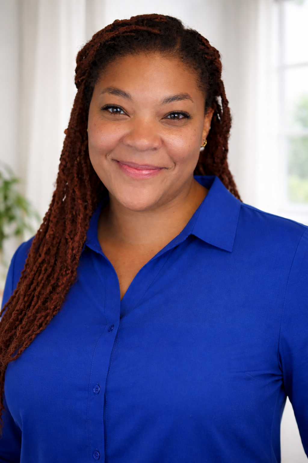 A woman with long reddish-brown dreadlocks smiling, wearing a royal blue collared shirt, with a background of a white wall and window.
