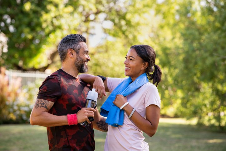 A man and woman smiling and talking outdoors in a park, dressed in workout clothes, with the man holding a water bottle.