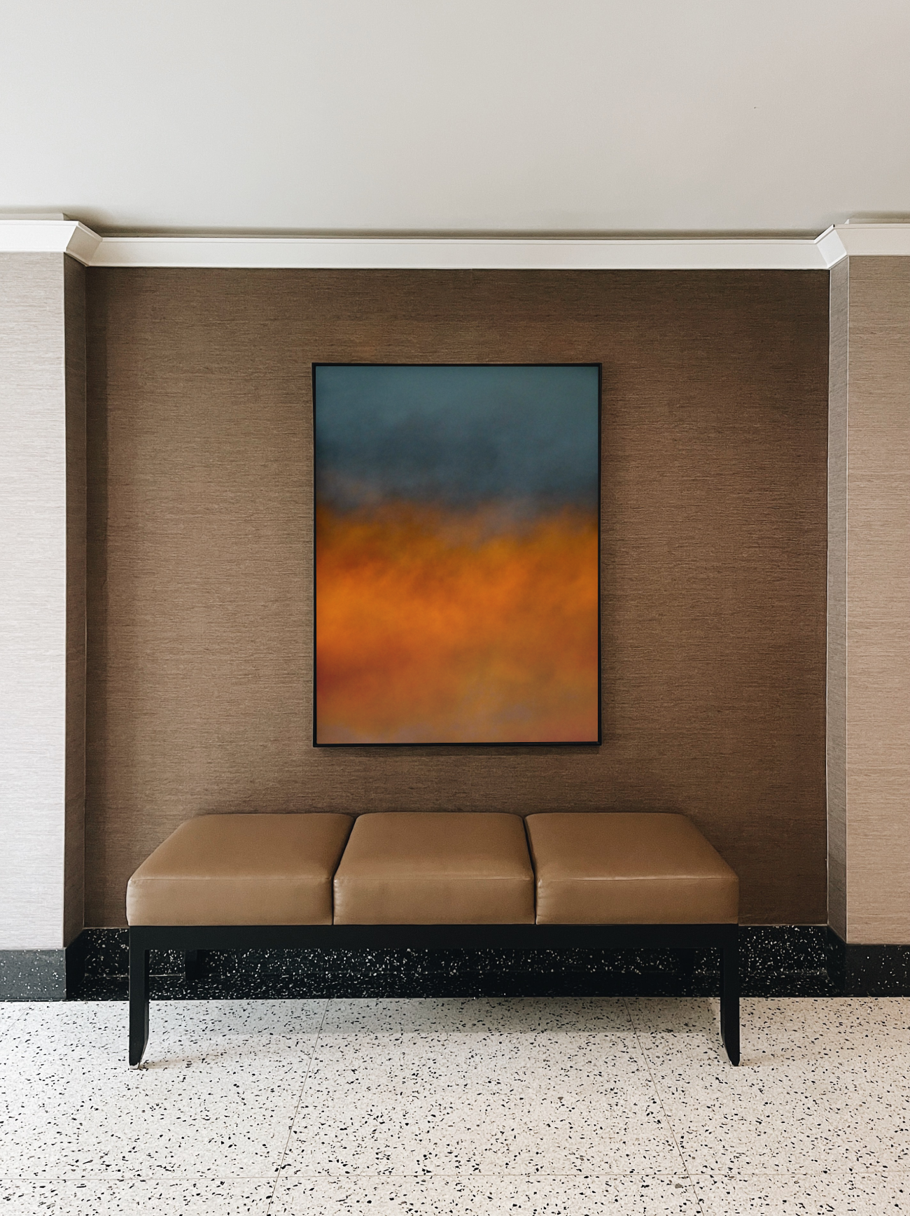 Ein Sitzbank vor einer braunen Wand, in einer hochwertigen Hotellobby, mit abstrakter Himmel-Fotografie in Blau, Orange und Gelb.