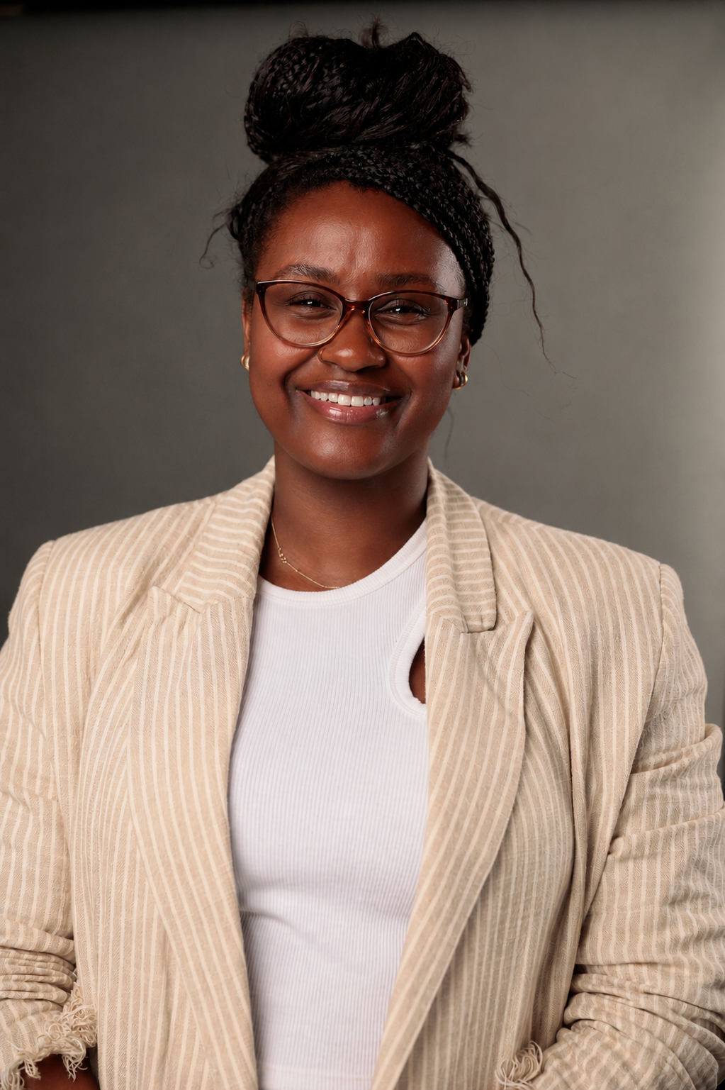 A smiling woman with glasses and braided hair styled in a bun, wearing a beige pinstripe blazer over a white top, standing against a dark gray background.