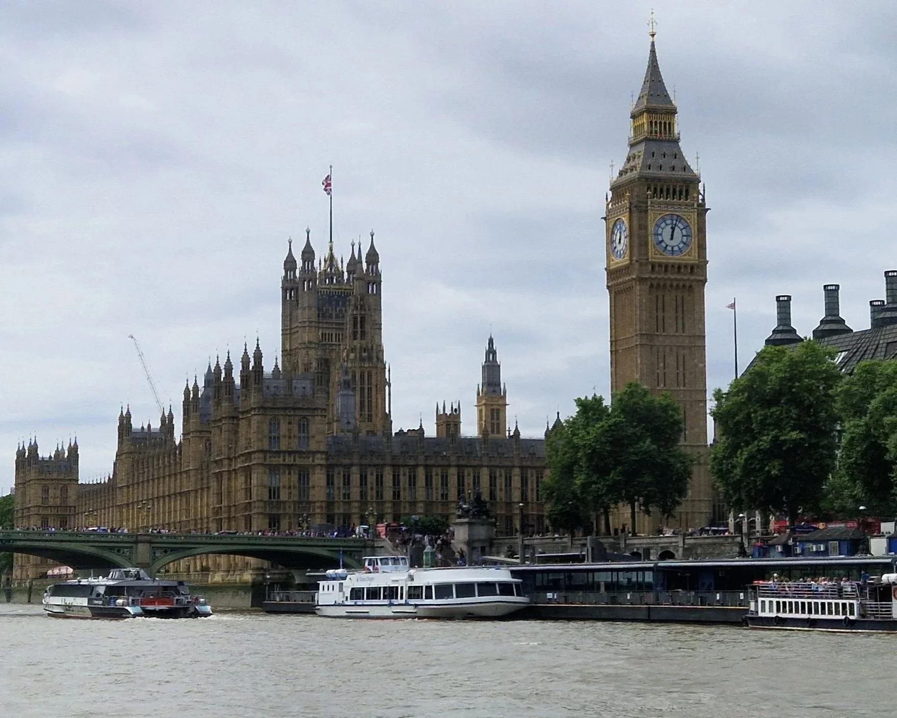 View of the Houses of Parliament and Big Ben in London, England, with boats on the River Thames and trees in the foreground.