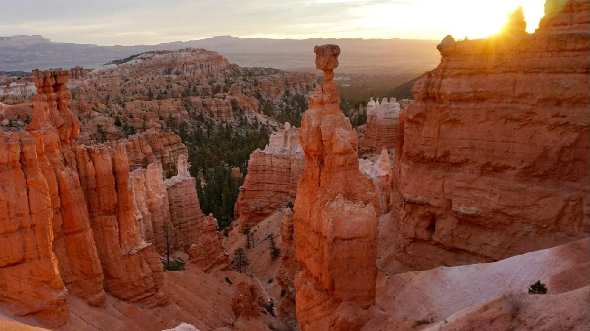 A canyon landscape with tall orange and white rock formations, some with mushroom-like shapes, at sunset with the sun setting behind distant mountains.