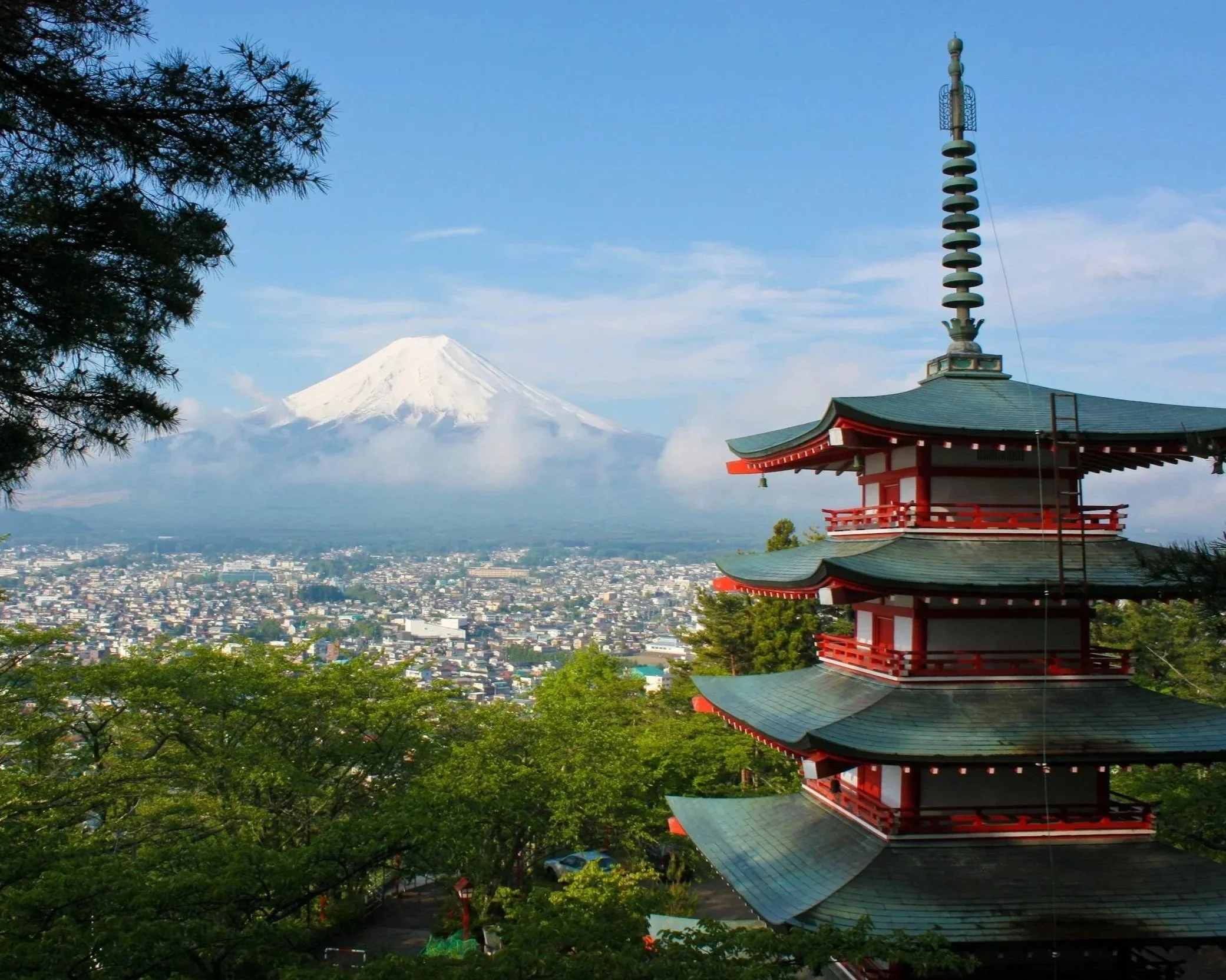 Traditional Japanese pagoda with red railings and green tiled roof, trees on the foreground, Mount Fuji with snow on top in the background, blue sky with some clouds.