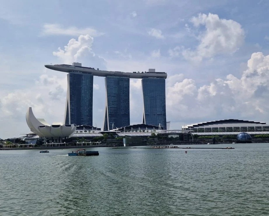 View of Marina Bay Sands resort in Singapore, featuring three tall hotel towers with a large rooftop structure resembling a ship, and an adjacent artscience museum shaped like a lotus flower, with water in the foreground and partly cloudy sky above.