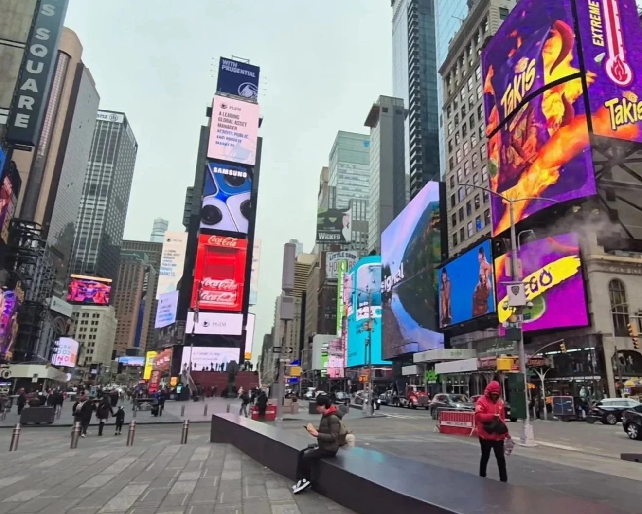 Times Square in New York City filled with bright digital billboards, some people walking and sitting, with tall buildings in the background on a cloudy day.
