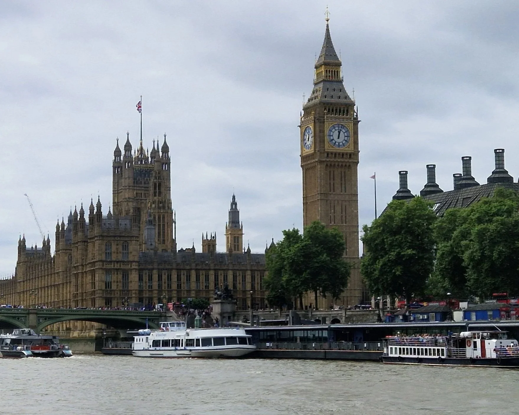 View of the Houses of Parliament and Big Ben clock tower in London, with boats on the River Thames and cloudy sky overhead.