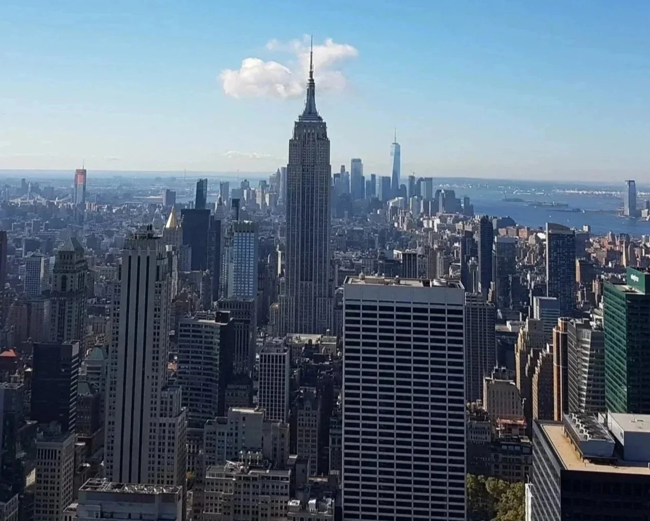 A view of New York City skyline featuring the Empire State Building in the foreground and One World Trade Center in the background with a blue sky and some scattered clouds.