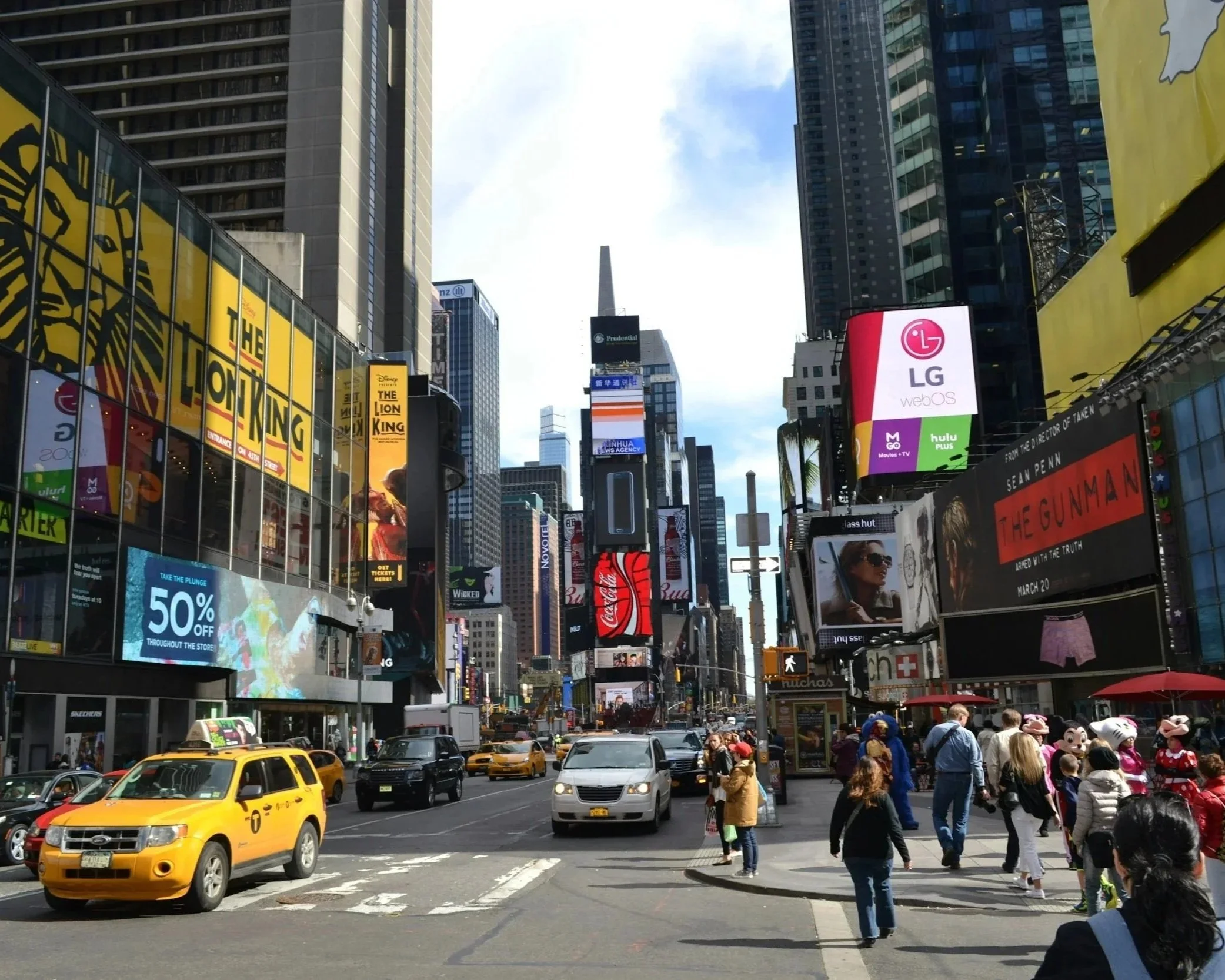 A busy city street scene in Times Square, New York City, with yellow taxis, pedestrians, and large digital billboards displaying advertisements and logos, including Disney's The Lion King and LG.