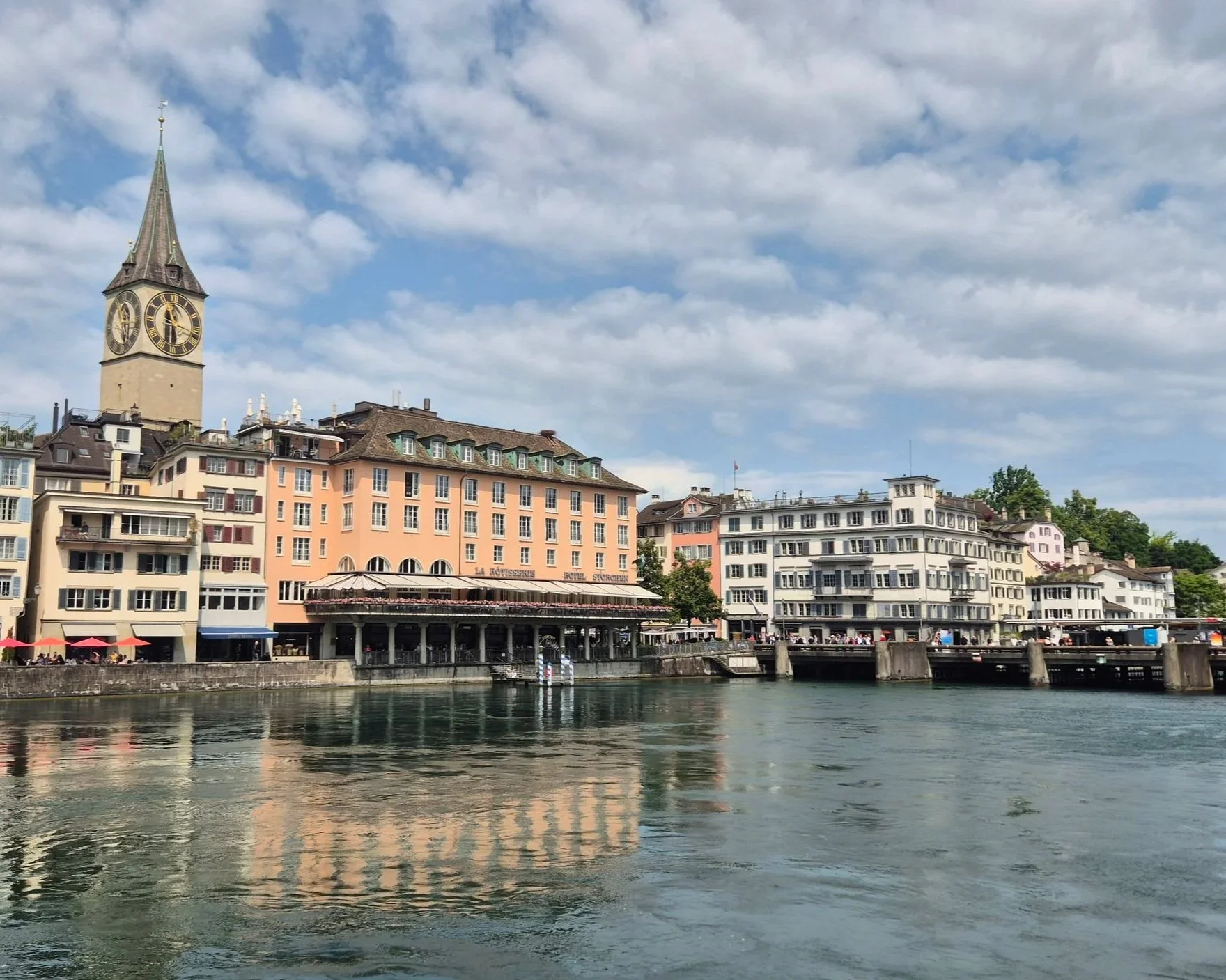 A scenic view of European-style buildings near a river, with a prominent clock tower on the left, under a cloudy sky.