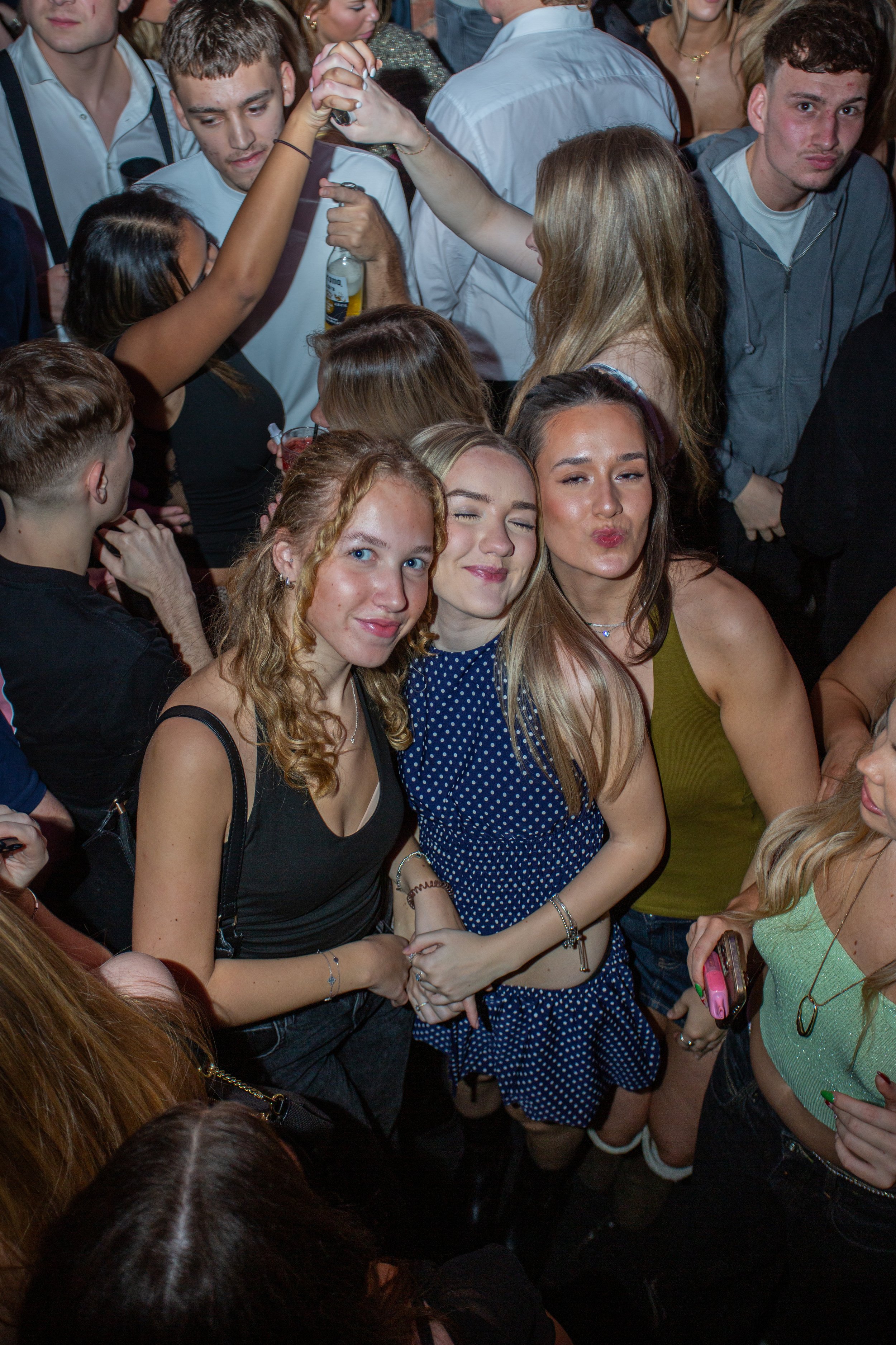 Group of people partying at a crowded event, with three young women smiling and making faces in the foreground.