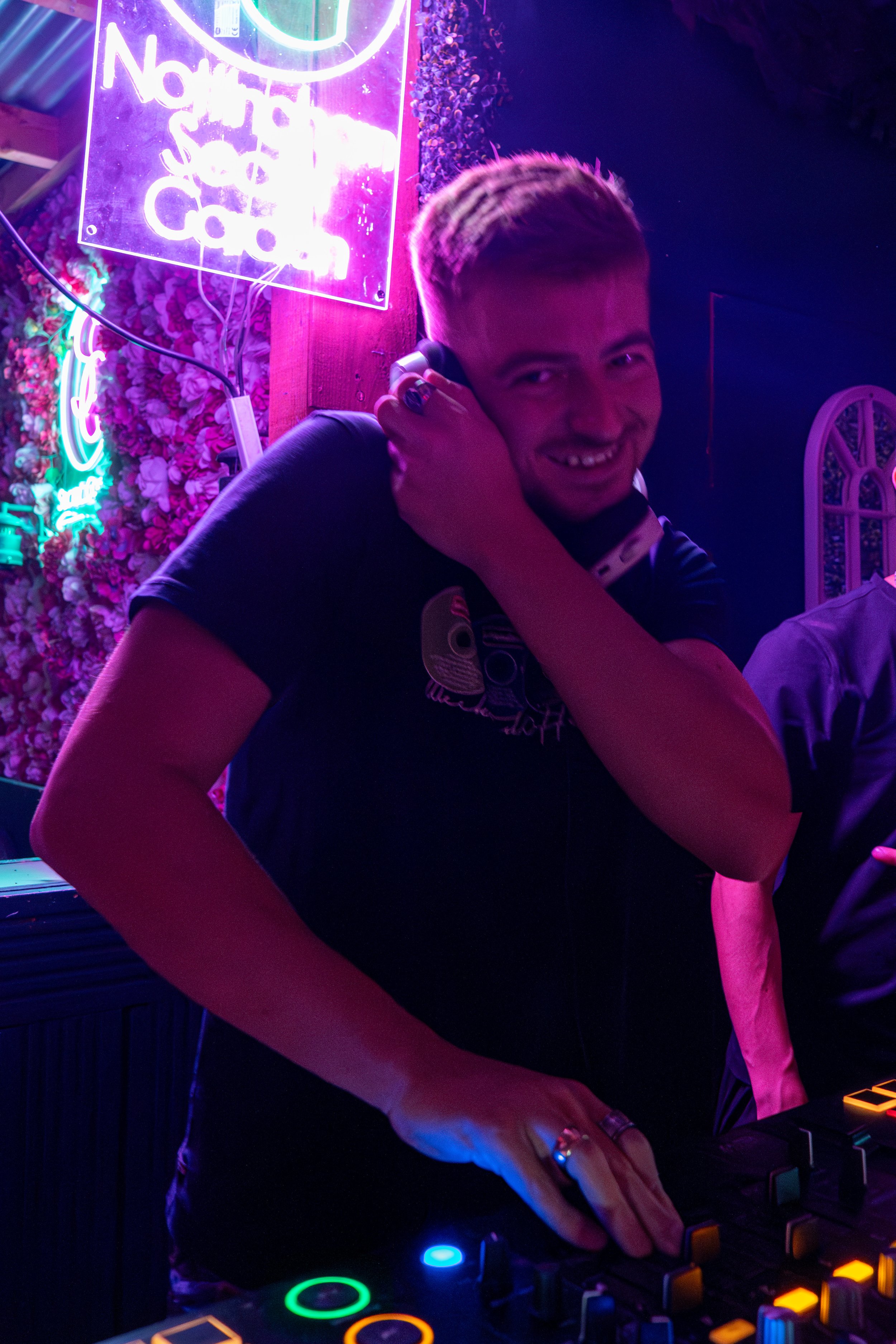 A young man with blonde hair, smiling, wearing a black shirt, holding headphones to his ear in a neon-lit nightclub with purple, pink, and blue lighting, a neon sign, and floral decorations.