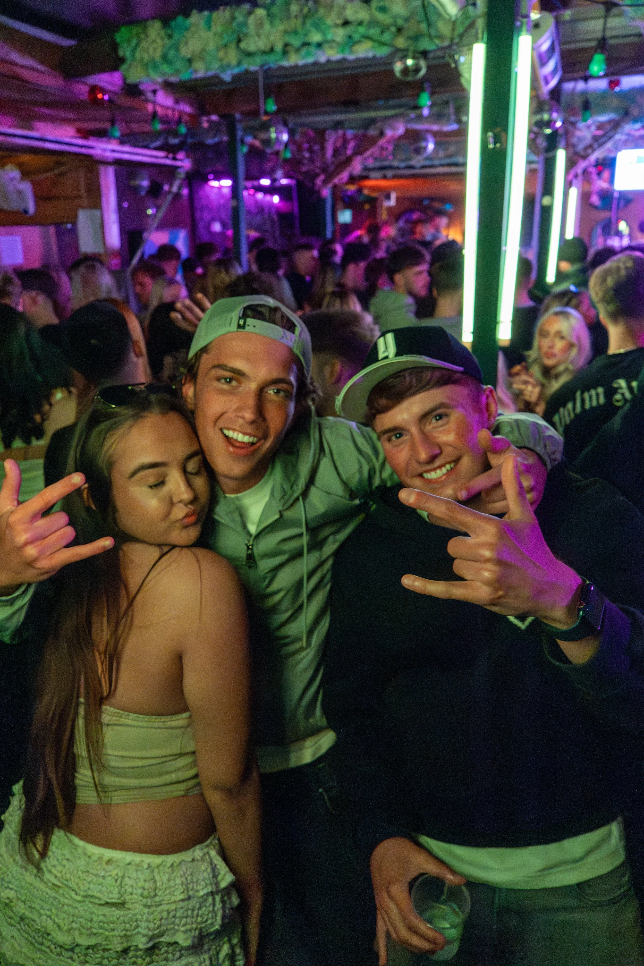 Three young people at a crowded nightclub, posing and smiling for the camera with purple and green lighting in the background.